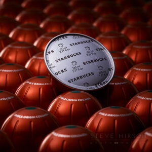Starbucks Nespresso Vertuo pods in Single-Origin Colombia flavor, featuring a close-up of one pod’s foil top with a sleek and textured background of bronze pods. Product photography by Steve Hirsch.