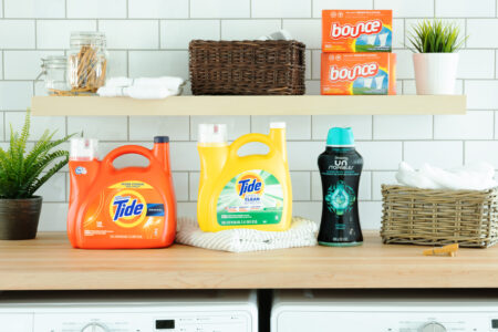 Laundry room setup featuring Tide Original and Tide Simply Clean & Fresh detergents, Bounce dryer sheets, and Downy Unstoppables scent booster, styled with wicker baskets, towels, and houseplants. Product photography by Steve Hirsch.